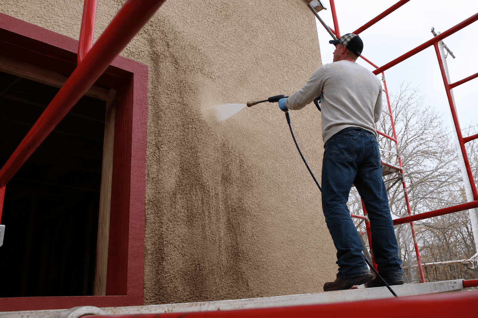 Worker pressure washing mildew and dirt from a textured stucco exterior wall while standing on scaffolding.